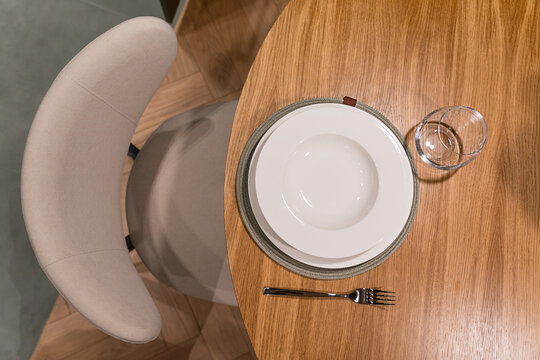 Top view of place setting of empty white round plate, drinking glass and silver fork  served on round wooden table at home kitchen with soft comfortable chair prepared for rustic lunch or dinner