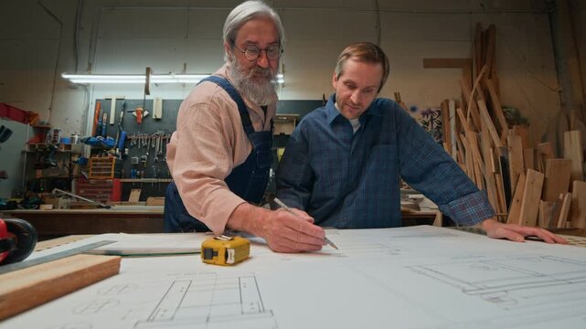 Senior Caucasian carpenter explaining blueprint schematics to middle aged apprentice in carpentry workshop. Clip suited for vocational training videos and craftsmanship advertising