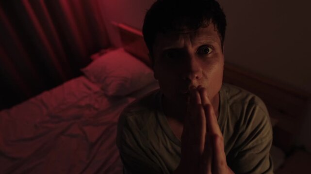 Desperate man praying with clasped hands and pleading directly to God, seeking mercy and forgiveness with an intense gaze in a dark bedroom with dramatic red low key lighting and deep shadows