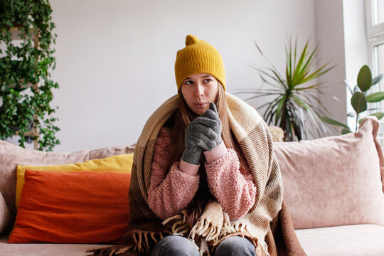 Woman bundled in warm clothes sitting on sofa and feeling cold indoors. Winter comfort and staying warm at home. The concept of a broken heating system or high cost of energy resources.