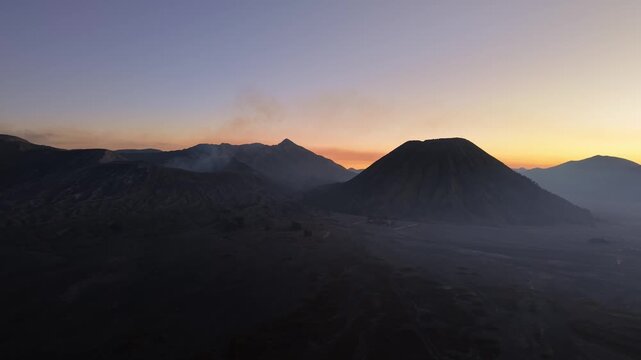 Mountains glow at dawn, smoke rises gently. Silhouettes stand stark against a soft, fading sky near Bromo