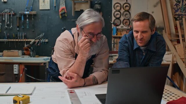 Senior Caucasian carpenter and middle aged Caucasian artisan discussing project on laptop in woodworking studio. Video ideal for craft education, trade school campaigns, joinery ads