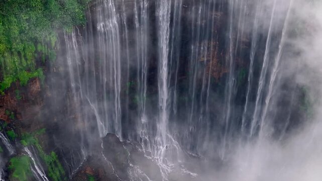 Powerful waterfalls cascade down steep rocky cliffs. Mist swirls around the falling water at Tumpak Sewu