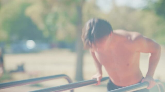 Athletic man performs calisthenics dips on parallel bars, then rests and catches his breath in a sunny outdoor park.