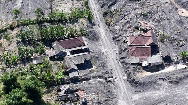 Drone aerial of lava scarred supiturang village showing destroyed roofs, ash covered terrain, debris, and surviving vegetation