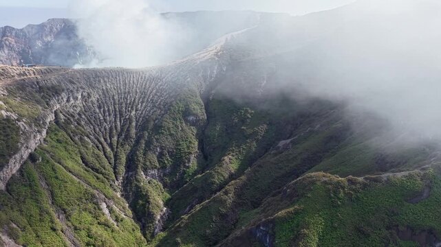 Misty clouds drift above volcanic caldera walls. Deep green slopes slope into rugged at Mount Ijen
