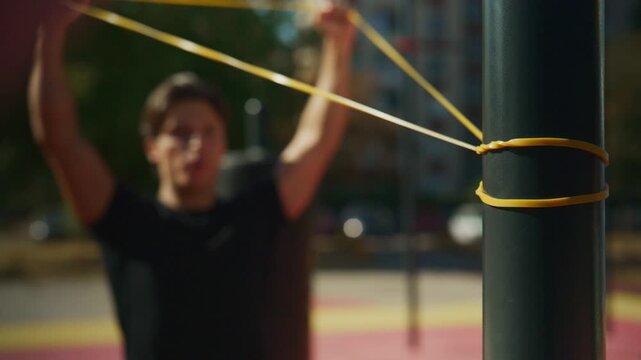 Young man performs resistance band training at an outdoor gym, pulling the band in controlled upper body exercise as focus shifts from the band to him.