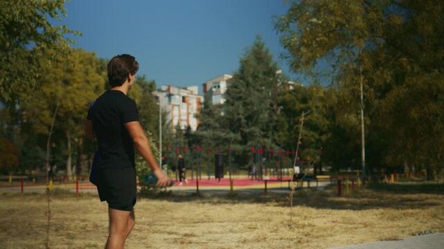 Fit man walks through a sunny park to an outdoor gym carrying a gym bag and water bottle for workout training.