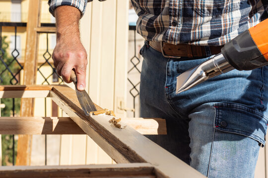 Carpenter using heat gun and wood scraper removes paint from old wooden window. Construction industry, carpentry. Restoration. 