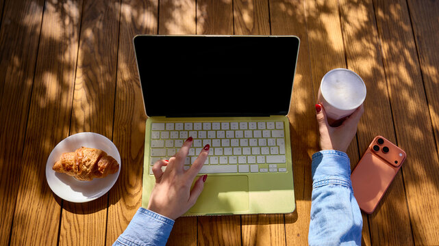 Female hands using laptop beside croissant and holding frothy coffee at sunlit wooden table, overhead view. Cafe workspaces, mobile connectivity and freelance lifestyle concept.
