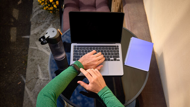 Man checking smartwatch beside laptop, water bottle and notebook on round outdoor table, overhead view. Time management, wearable technology and flexible employment concept.
