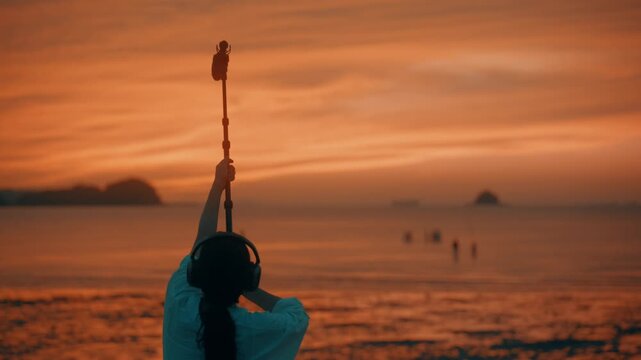 Rear View of Woman Wearing Headphones and Raising Shotgun Microphone High on Boom Pole at Sunset Beach