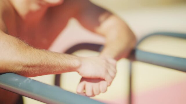 Athletic man rests on dip bars, catches his breath, and stretches his wrists after an outdoor calisthenics workout in slow motion.