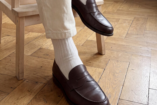 Closeup view of man in dark leather loafers, white ribbed socks and light trousers sits on light chair in apartment during day. One foot rests on chair rung while other stands on wooden floor