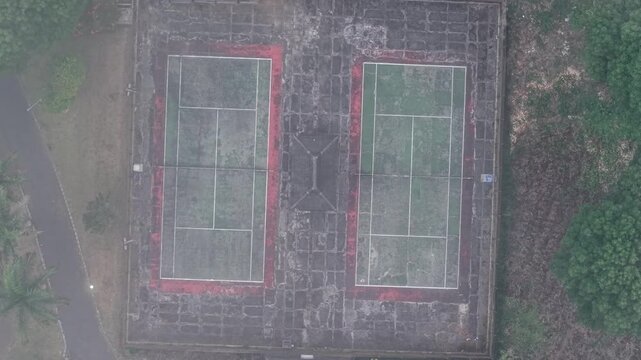 Aerial view shows two weathered tennis courts. Red lines contrast with faded green surfaces