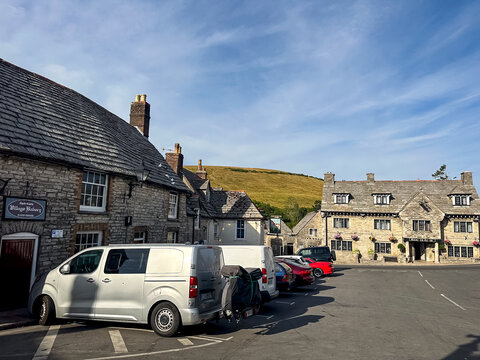 Corfe Castle, Dorset, UK - July 13, 2026. Village Square Square with Bakery and Bankes Arms Hotel in Corfe Castle. A high-detail horizontal landscape photograph showing parked cars in the main village