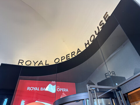 London, UK - July 23, 2026. Entrance Sign and Glass Facade of the Royal Opera House in London. A high-detail horizontal low-angle photograph capturing the modern curved glass entrance and metallic let