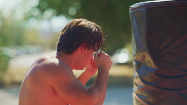 Shirtless man throws jab and cross combinations on a punching bag outdoors, then pauses exhausted after intense boxing training in slow motion.