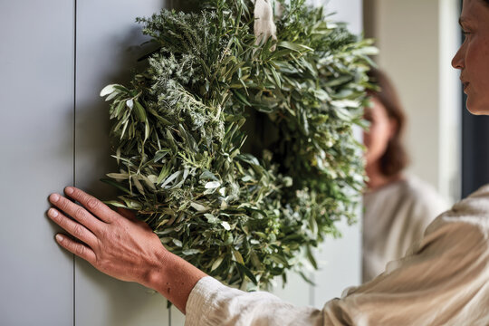 A person gently adjusting a carefully crafted green wreath hanging on a front door, symbolizing the welcoming nature of home and the beauty of seasonal decorations.