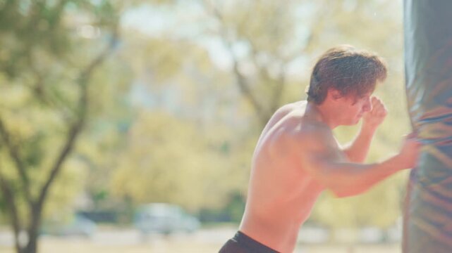 Young adult man punches a heavy bag outdoors, then catches his breath and wipes sweat after fitness training in a park.