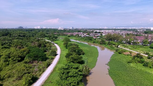 Muddy river winds through green fields and trees. Sky clear blue. Paths run alongside water at Kampung Terus