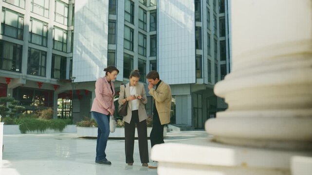 Three business colleagues gather outdoors, focused on a smartphone screen. They are engaged in a shared digital experience, reviewing information together.