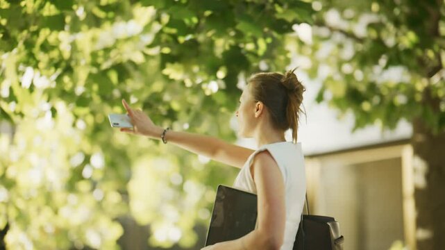A confident young woman walks through a green urban environment, carrying a stylish bag and tablet. She navigates the city park on a beautiful, sunny day.
