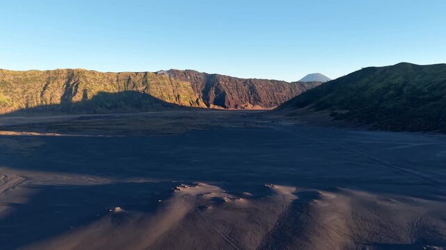 Golden light bathes volcanic crater floor. Dark sand dunes stretch across rugged landscape at Sea of Sand Bromo