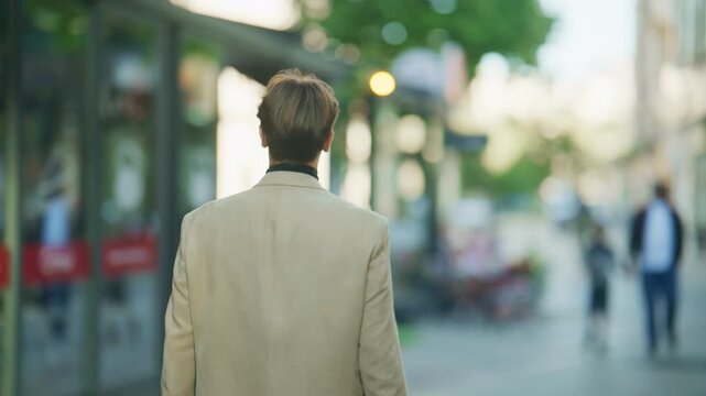 A stylish man in a beige blazer walks away from the camera on a vibrant city street. He is seen moving through a busy urban area during the daytime.