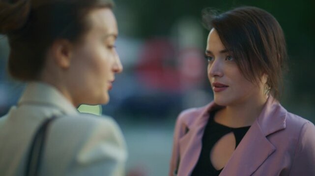Two young women engage in a pleasant conversation outdoors, sharing smiles and gestures. They are seen enjoying each other's company during a casual meeting.