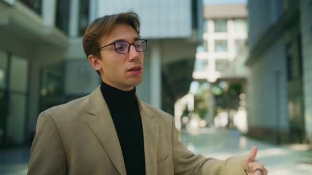 A stylish young man in glasses and a blazer actively discusses a topic with a colleague outdoors. He gesticulates confidently while having an engaging conversation in an urban setting.