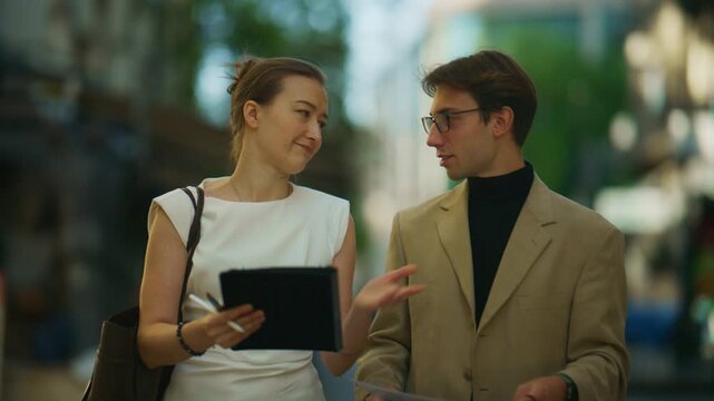 Two business professionals walk together, reviewing information on a tablet in an urban setting. They engage in a dynamic discussion, collaborating on their shared work project.