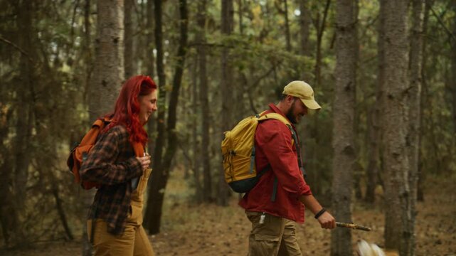 Couple carries backpacks with their dog through a sunny autumn forest while the man playfully holds a stick.