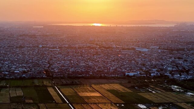 Golden sun dips over sprawling city and fields. City sprawls below, bathed in soft orange glow at Surabaya