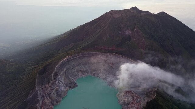 Aerial view of a volcanic crater with mist rising. Turquoise water contrasts with dark, rugged rock at Mount Ijen