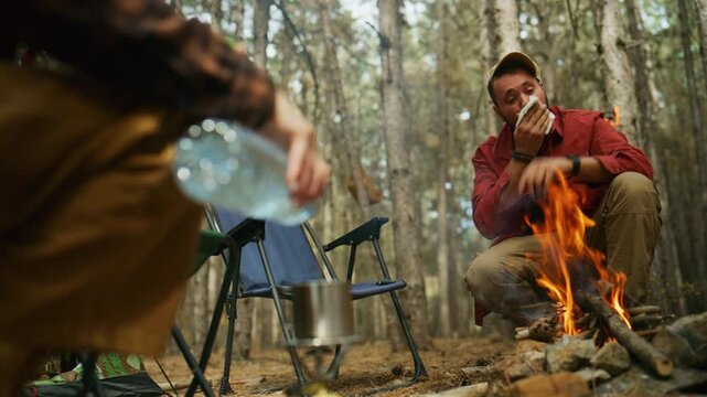 Camper pours water into a mug on a portable stove while another man rests beside a campfire in the forest.