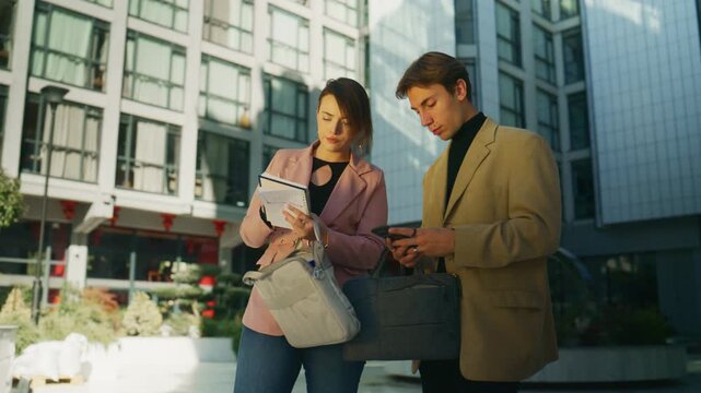 Two business professionals collaborate outdoors, reviewing notes and using a smartphone. They are discussing information in a modern urban setting.