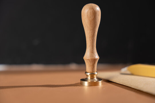 Wooden wax seal stamp with a brass head stands on a brown surface beside a kraft envelope and a gold wax stick against a dark background.