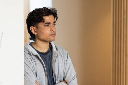 Man leaning against white partition, facing right arms crossed, in gray hoodie at home, copy space