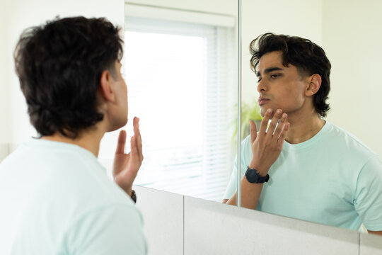 Man examining face and touching chin at bathroom vanity with mirror, blinds, plant and watch