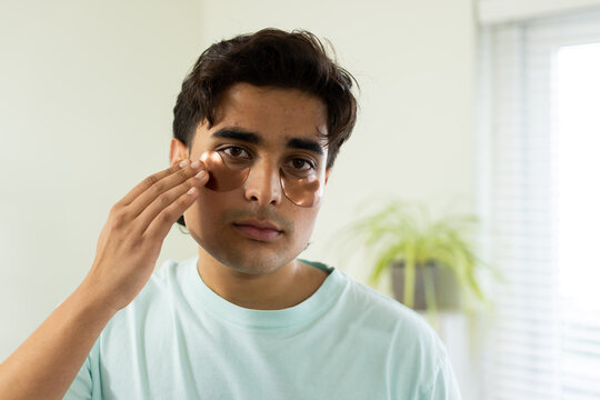 Indian man applying bronze eye patches while wearing mint T-shirt at home near window and plant