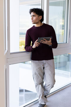 Asian man leaning on window ledge holding tablet, wearing burgundy sweater and gray cargo pants