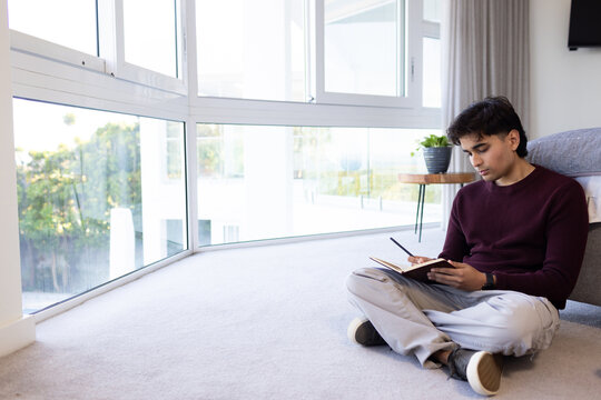 Asian man sitting cross-legged scribbling in notebook with pen at home by curved windows