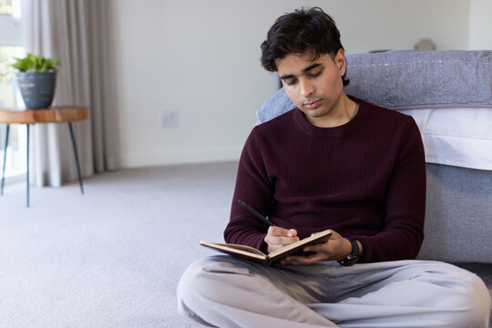 Indian man sitting on bedroom carpet, writing in notebook wearing burgundy sweater, copy space