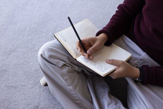Adult male in burgundy sweater cross-legged on gray carpet writing on lined notebook with black pen