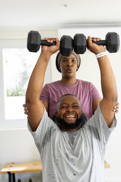 Couple pressing hex dumbbells overhead, woman standing behind spotting, wearing mauve tee at home
