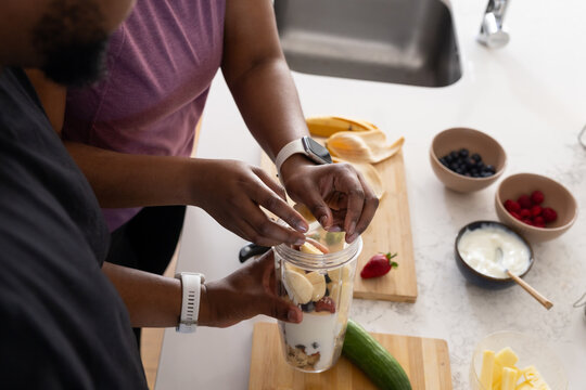 Couple making yogurt smoothie at countertop adding banana to clear blender cup with wristbands
