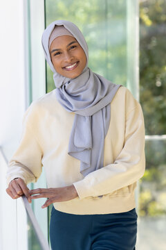 African American woman wearing hijab and cream top standing by railing in atrium with glass panels
