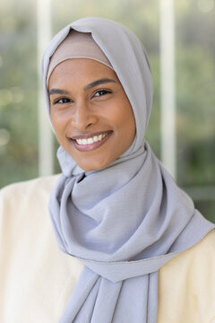 Woman smiling, facing camera at home in window light, wearing gray hijab beige undercap and top