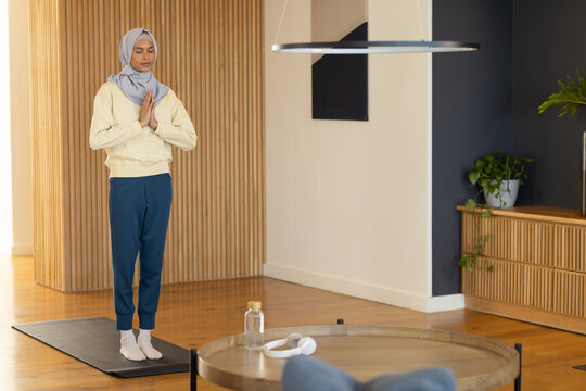 Woman meditating on black mat at home wearing headscarf holding palms near water bottle